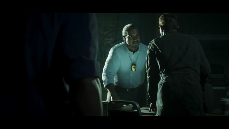 Méndez, a man in a white shirt with a gold badge on a chain, leans over a desk towards a man in dark overalls in a dim room.
