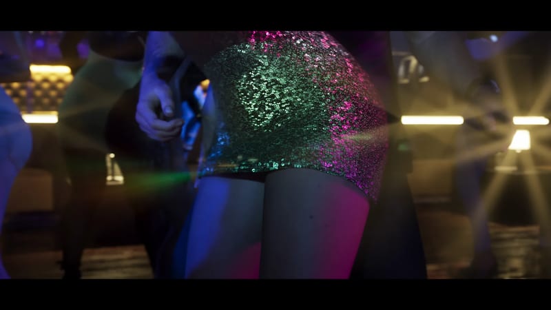 A woman in a short green sequinned dress dances in a crowded, dimly lit nightclub lit by pink, blue and yellow lights.