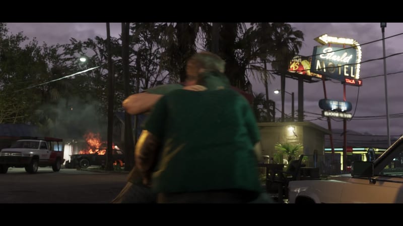 Two men embrace in a motel car park at dusk, seen from behind, with the Starlet Motel sign glowing and a car burning in the background.