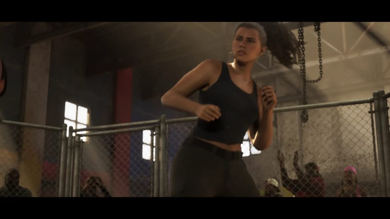 Lucia Caminos, in a black tank top and dark trousers, moves in a fighting stance inside a fenced ring in a dusty warehouse gym with spectators behind.