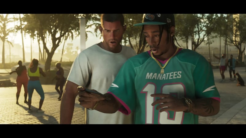Jason Duval and a man in a teal Manatees jersey look at a phone on a sunny, palm-lined paved plaza, with people exercising behind them.