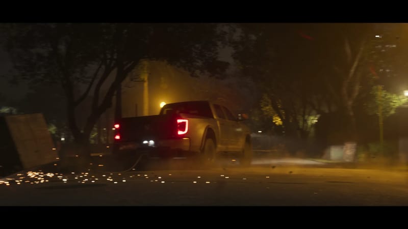 A pale pickup truck with red tail lights drags a cable along a dark, tree-lined street at night, throwing up sparks and dust.