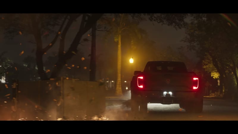 A dark pickup truck with glowing red tail lights drives away along a leafy, lamp-lit street at night, with leaves blowing through the air.