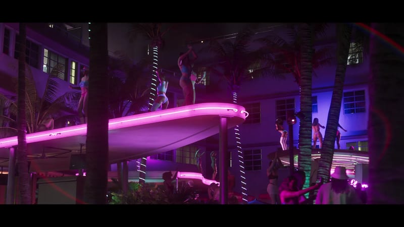 Dancers in swimwear perform on a curved, pink neon-lit platform among palm trees outside a hotel at night, with a crowd below.