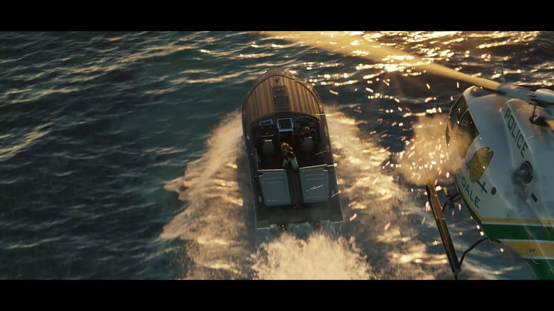 A police helicopter chases a wooden speedboat with two people aboard across choppy sea at golden hour, sparks flying near the helicopter.