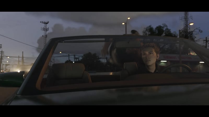 A young woman with short curly hair sits at the wheel of a dark convertible on a road at dusk, with street lights and a radio mast behind.