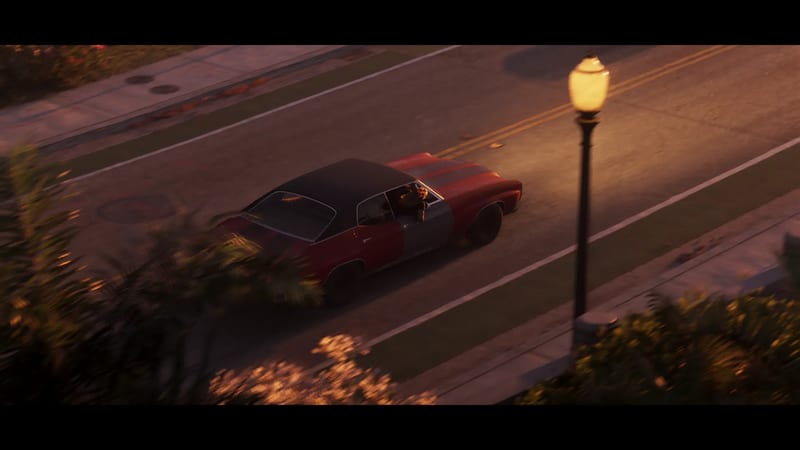 A red car with a black roof and a grey door drives along a road at dusk, seen from above, with a person leaning out of the window and a lit street lamp beside it.