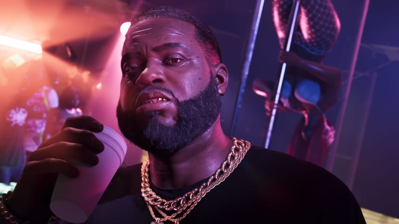 Boobie Ike, a bearded man in gold chains, holds a white cup in a club with pink and orange lighting and a dancer on a pole behind him.