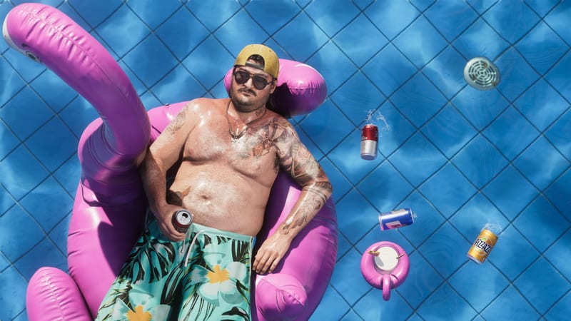 Cal Hampton, a tattooed man in sunglasses and floral swim shorts, lounges on a pink inflatable flamingo in a tiled blue swimming pool, holding a can.