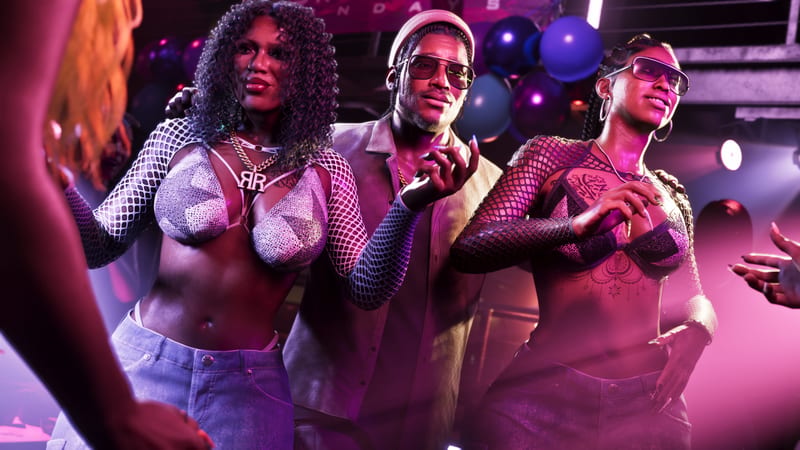 Three people pose together in a pink and purple lit nightclub, a man in sunglasses and a beanie between two women in fishnet tops.