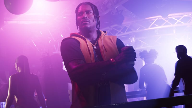 Dre'Quan Priest stands with arms folded, holding a cup, in a hazy purple-lit nightclub with other clubgoers behind him.