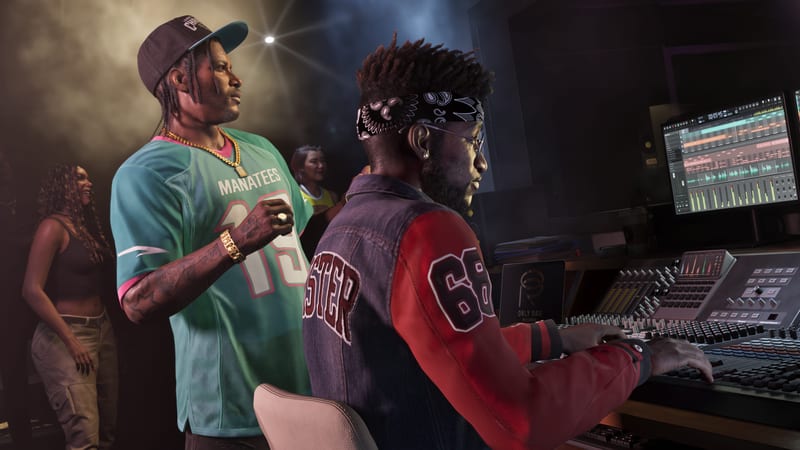 A man in a teal Manatees jersey stands beside a seated man in a bandana operating a mixing desk in a recording studio.