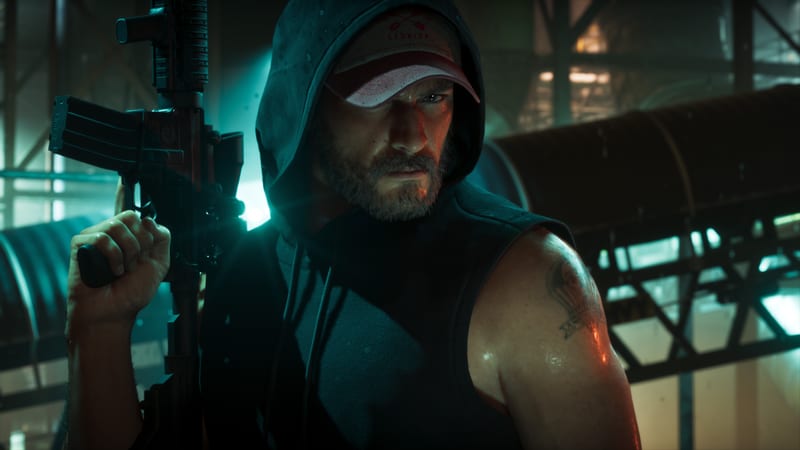 Jason Duval, bearded and wearing a dark hooded sleeveless top and a cap, holds a rifle upright in a dim industrial space lit with teal light.