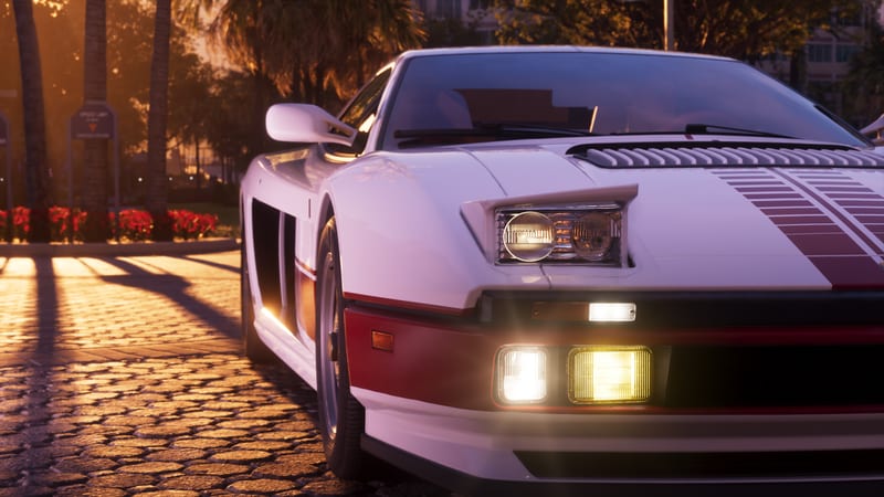 The front of a white and red '95 Grotti Cheetah with its lights on, parked on cobbles at sunset beside palm trees and red flowers.