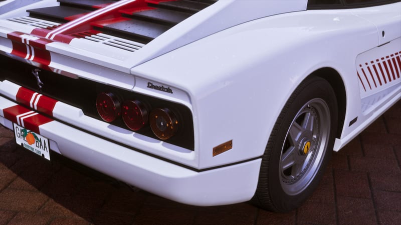 Rear corner of a white '95 Grotti Cheetah with red stripes, showing its triple tail lights, alloy wheel and a peach-themed number plate, on red brick paving.