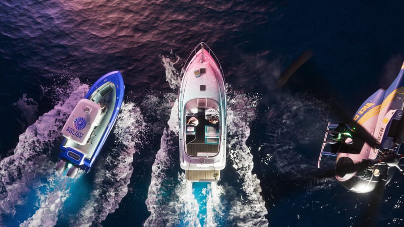 Aerial view of a pink and white Shitzu Squalo speedboat racing across dark water, chased by a Vice City Police boat and a police helicopter.