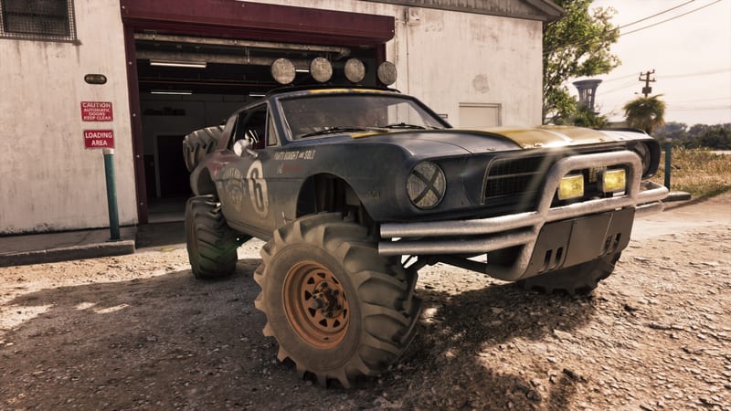A dirty dark blue '67 Vapid Dominator Buggy with huge tyres and roof lights stands outside a garage on a gravel yard in daylight.