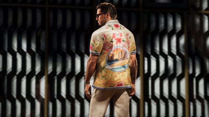 A man in sunglasses, seen from behind, wears a cream shirt printed with flamingos, hibiscus flowers and a beach skyline, in front of a dark patterned screen.
