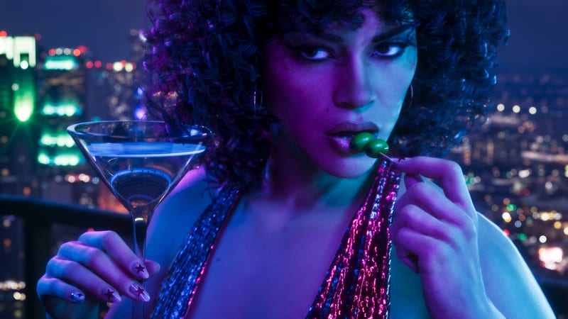 A woman with curly dark hair eats an olive from a cocktail stick, holding a martini glass on a balcony above a city lit up at night.